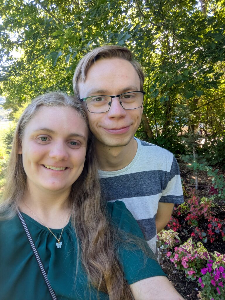 Brooke standing in a selfie with Jon over her shoulder, flowers and bushes next to them.